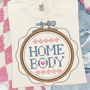 May include: Cream-coloured t-shirt with a cross-stitch design. The design features the words "HOME BODY" in blue, a pink heart, and a brown embroidery hoop. The shirt is made of a soft material and is perfect for casual wear.