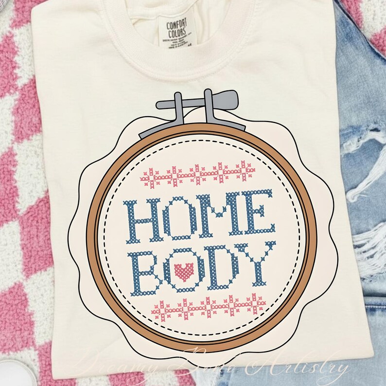 May include: Cream-coloured t-shirt with a cross-stitch design. The design features the words "HOME BODY" in blue, a pink heart, and a brown embroidery hoop. The shirt is made of a soft material and is perfect for casual wear.
