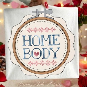 May include: A white fabric with a cross-stitch design in a wooden embroidery hoop. The design features the words "HOME BODY" in blue, with a pink heart. The top and bottom of the design have a decorative border. The text "Dreamy Boho Artistry" is at the bottom.