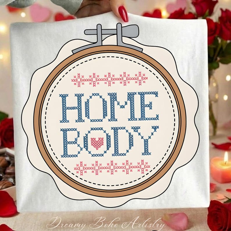 May include: A white fabric with a cross-stitch design in a wooden embroidery hoop. The design features the words "HOME BODY" in blue, with a pink heart. The top and bottom of the design have a decorative border. The text "Dreamy Boho Artistry" is at the bottom.