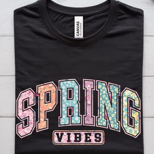 May include: Black t-shirt with the words "SPRING VIBES" in a floral design. The word "SPRING" is in a bold, block letter style with a floral pattern in pink, orange, blue, and green. The word "VIBES" is in a rectangular box.