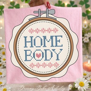 May include: A pink fabric with a cross-stitch design in a wooden embroidery hoop. The design features the words "HOME BODY" in blue, with a pink heart. The top and bottom of the design have a repeating pattern. The text "Dreamy Boho Artistry" is at the bottom.