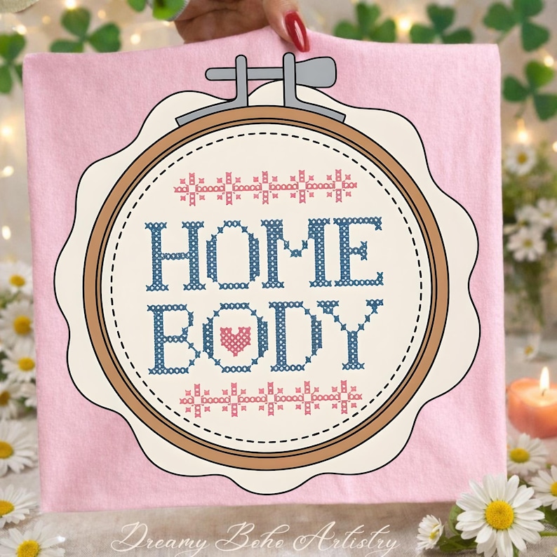 May include: A pink fabric with a cross-stitch design in a wooden embroidery hoop. The design features the words "HOME BODY" in blue, with a pink heart. The top and bottom of the design have a repeating pattern. The text "Dreamy Boho Artistry" is at the bottom.