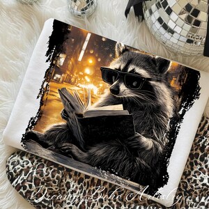 May include: White sweatshirt featuring a raccoon wearing sunglasses and reading a book. The raccoon is set against a city backdrop with warm lighting. The sweatshirt has a distressed black border and the text "Dreamy Boho Artistry".
