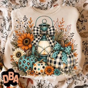 May include: A beige sweatshirt featuring a graphic print of a teal lantern with a black and white buffalo check bow, two pumpkins with teal leopard and buffalo check patterns, teal polka dot bows, and orange and yellow sunflowers.
