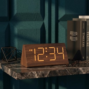 May include: A wooden digital alarm clock displaying 12:34 AM in amber numerals. The clock has a triangular shape and sits on a marble shelf. Books and a geometric metal sculpture are in the background.