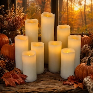 May include: A collection of nine cylindrical, off-white LED pillar candles of varying heights, each emitting a warm, soft glow. The candles are arranged on a dark, weathered wooden surface alongside several orange pumpkins and dried leaves.