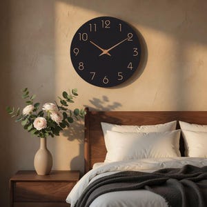 May include: A black round wall clock with gold-colored hands and numerals. The clock is mounted on a beige wall above a wooden nightstand with a vase of flowers and a bed with white pillows and a gray blanket.