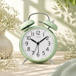 Puede incluir: Un reloj despertador analógico verde menta con esfera blanca y números negros. El reloj tiene un asa y patas de metal, y dos campanas en la parte superior. La palabra "PEAKEEP" está impresa en la esfera. El reloj está sobre un fondo suave.