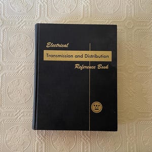 May include: A black hardcover book titled Electrical Transmission and Distribution Reference Book with gold lettering. A horizontal gold bar highlights the title, and a circular logo with a stylized W symbol is positioned in the lower right corner.