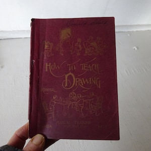 Peut inclure: Un livre bordeaux intitul&eacute; "How to Teach Drawing" de "Cooper" avec des illustrations d'enfants dessinant. Le livre est un manuel de formation au dessin.