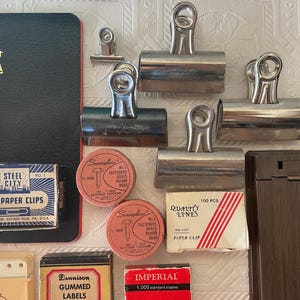 May include: An assortment of vintage office supplies, including metal binder clips, boxes of paper clips, labels, and fasteners. A black notebook with a gold bird emblem is also present. The items are arranged on a textured white surface.