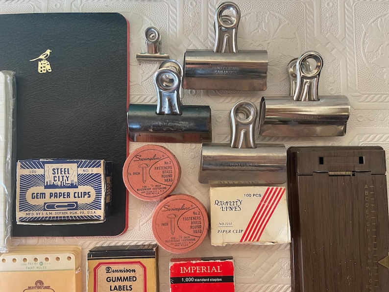 May include: An assortment of vintage office supplies, including metal binder clips, boxes of paper clips, labels, and fasteners. A black notebook with a gold bird emblem is also present. The items are arranged on a textured white surface.