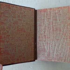 May include: An open book with a brown leather cover and red patterned endpapers. The endpapers feature a repeating design of various small objects, including tools, buildings, and plants.