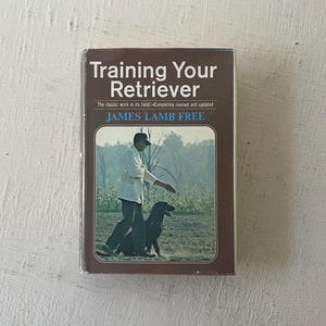 May include: A brown book titled "Training Your Retriever" by James Lamb Free. The cover features a black and white photo of a man training a black Labrador Retriever.
