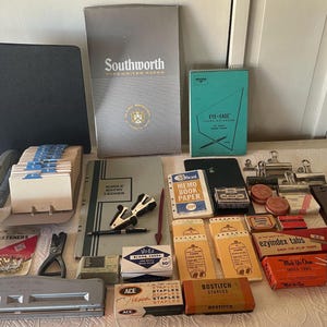 May include: A collection of vintage office supplies, including staplers, notebooks, index cards, and a hole punch. The items are in various colors, such as black, gray, and teal, and are arranged on a white surface. The image showcases a variety of stationery.