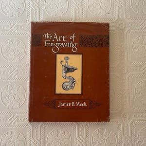 May include: A brown hardcover book titled The Art of Engraving by James B. Meek. The front cover features a central rectangular illustration of an ornate, metallic, scroll-shaped object with intricate detailing and a dark, decorative border.