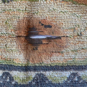 May include: Close-up of a beige, green, and brown rug with a tear in the center. The tear is repaired with a white thread.