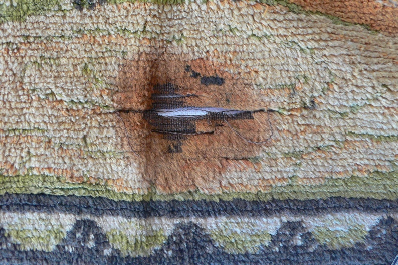 May include: Close-up of a beige, green, and brown rug with a tear in the center. The tear is repaired with a white thread.