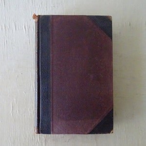 May include: A vintage hardcover book with a burgundy cover and black spine. The corners are reinforced with black material. The book's edges show signs of wear, suggesting its age and use. The overall aesthetic is classic and antique.