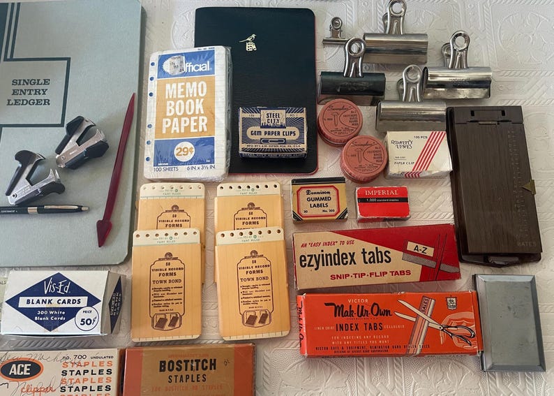 May include: An assortment of vintage office supplies, including a ledger, memo book paper, staplers, paper clips, index tabs, and blank cards. The items are in various colors, with visible text on the packaging.