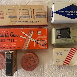 May include: A collection of vintage office supplies, including index tabs, labels, paper clips, and fasteners. The items are in their original packaging, with text and graphics. The color palette includes orange, beige, and white.