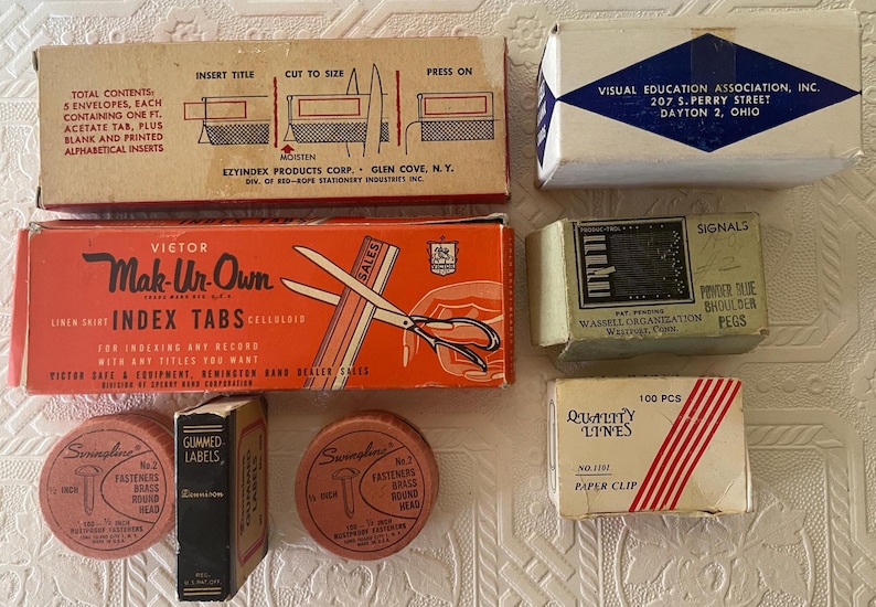 May include: A collection of vintage office supplies, including index tabs, labels, paper clips, and fasteners. The items are in their original packaging, with text and graphics. The color palette includes orange, beige, and white.