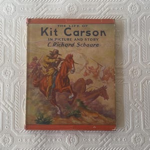 May include: A vintage book cover with a red background and the title "The Life of Kit Carson in Picture and Story" by C. Richard Schaare. The cover features an illustration of a group of cowboys on horseback riding through a landscape.