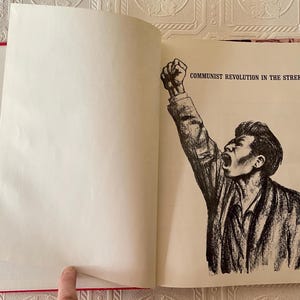 Puede incluir: Un libro abierto con lomo rojo y p&aacute;ginas blancas. La p&aacute;gina derecha presenta una ilustraci&oacute;n en blanco y negro de un hombre con el pu&ntilde;o en alto y el texto "COMMUNIST REVOLUTION IN THE STREETS".