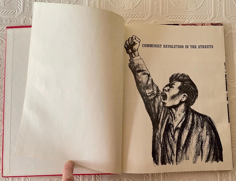 Puede incluir: Un libro abierto con lomo rojo y p&aacute;ginas blancas. La p&aacute;gina derecha presenta una ilustraci&oacute;n en blanco y negro de un hombre con el pu&ntilde;o en alto y el texto "COMMUNIST REVOLUTION IN THE STREETS".