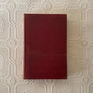 May include: A vintage, burgundy-colored book with a debossed emblem on the cover. The book has a rectangular shape with a slightly worn appearance, suggesting age. The cover is a solid, deep red hue, and the edges are visible.