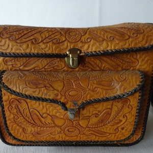 May include: A brown leather satchel bag with intricate embossed floral designs. The bag has two flap closures with brass buckles and a brown adjustable shoulder strap.