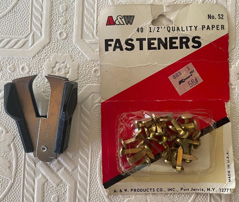 May include: An image of office supplies. A package of brass fasteners and a black and silver staple remover are displayed. The fastener package has red and white packaging with the text "FASTENERS". The staple remover is made of metal and black plastic.