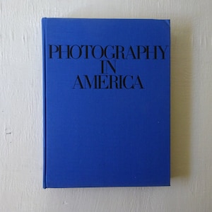 May include: A blue hardcover book with the title "Photography in America" printed in black.