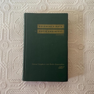 May include: A dark green book titled "REFERENCE DATA for RADIO ENGINEERS" with gold lettering. The book is a second edition, published by Federal Telephone and Radio Corporation. The book is on a patterned white surface.