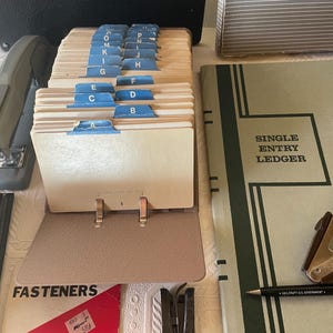 May include: An assortment of vintage office supplies, including a card file with blue tab dividers labeled A through P, a stapler, a ledger with "SINGLE ENTRY LEDGER" text, and a pen. The items are arranged on a light-colored surface.