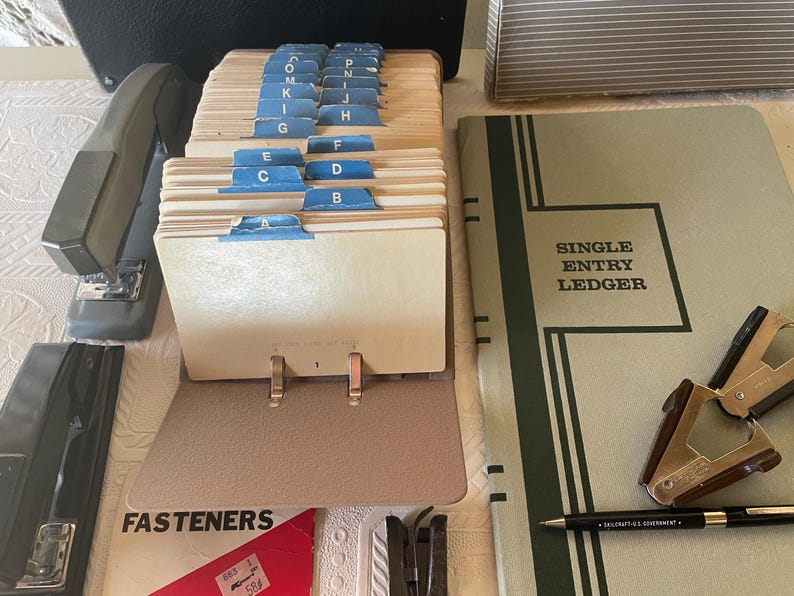 May include: An assortment of vintage office supplies, including a card file with blue tab dividers labeled A through P, a stapler, a ledger with "SINGLE ENTRY LEDGER" text, and a pen. The items are arranged on a light-colored surface.