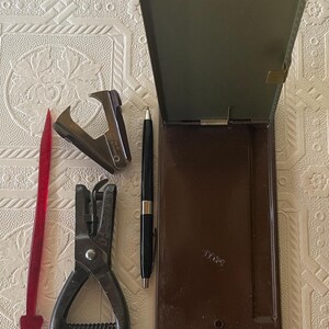 May include: An assortment of vintage office supplies, including a stapler, hole punch, pen, and a red plastic letter opener. A brown hinged box is also present. The items are arranged on a textured, off-white surface.