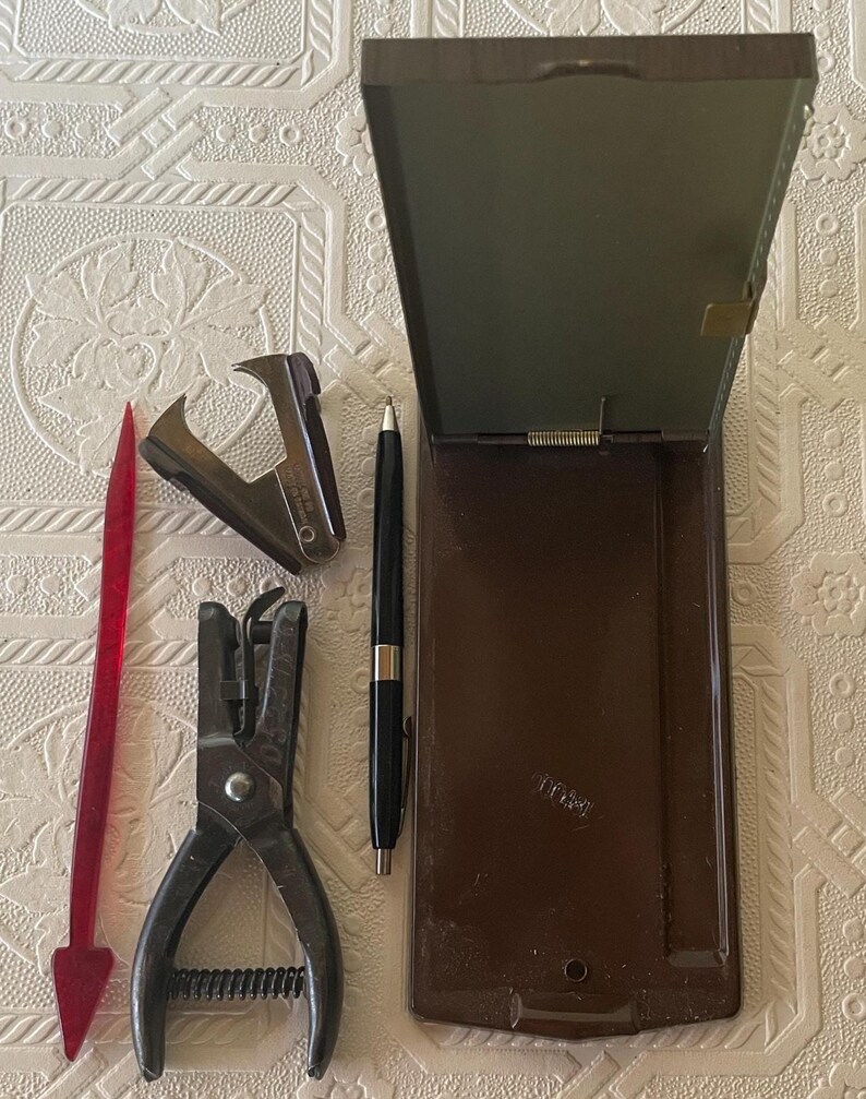 May include: An assortment of vintage office supplies, including a stapler, hole punch, pen, and a red plastic letter opener. A brown hinged box is also present. The items are arranged on a textured, off-white surface.