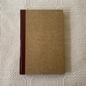 May include: A hardcover book with a textured, tan cover and a dark burgundy spine. The book is rectangular and appears to be closed. The background is a white, patterned surface.