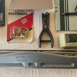 May include: An assortment of vintage office supplies, including a stapler, hole punch, fasteners, a pen, and a paper punch. The items are arranged on a textured surface, showcasing a collection of tools for paper organization and office work.