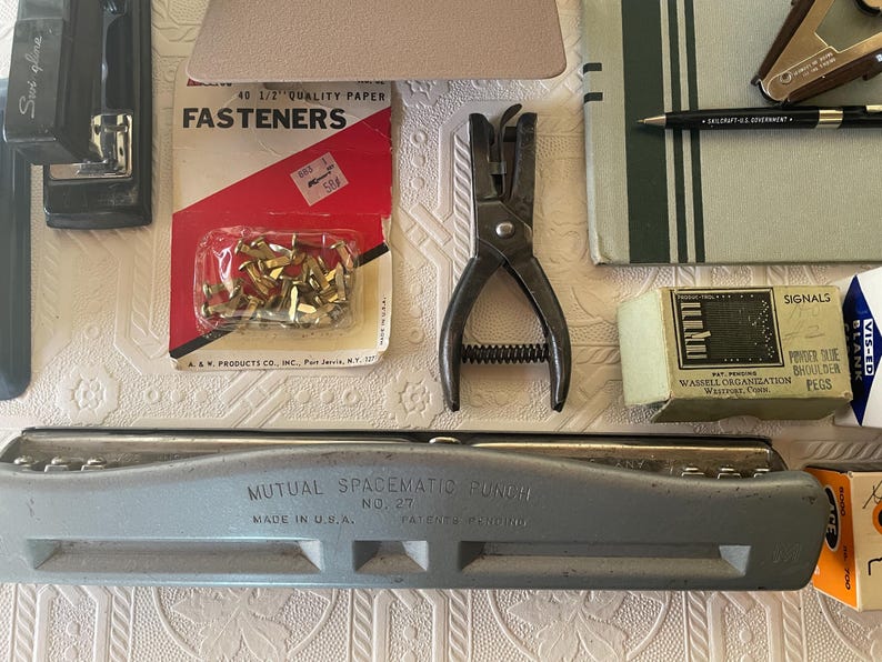 May include: An assortment of vintage office supplies, including a stapler, hole punch, fasteners, a pen, and a paper punch. The items are arranged on a textured surface, showcasing a collection of tools for paper organization and office work.