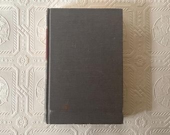May include: A hardcover book with a dark gray cover. The spine is a darker shade of gray with a red accent. The book is closed and resting on a patterned surface. The book appears to be vintage or antique.