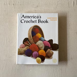 May include: A book titled "America's Crochet Book" by Gertrude Taylor. The cover features a basket overflowing with colorful yarn balls.