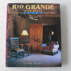 May include: Book cover featuring a rustic interior with wooden furniture, a lamp, and a rug. The title is "Rio Grande High Style Furniture Craftsmen" by Elmo Baca.