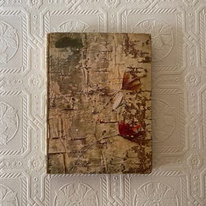 May include: A rectangular book with a weathered, textured cover featuring a distressed beige and brown finish. Two delicate, dried floral stems are attached to the front cover, one with a pale petal and one with a deep red, textured bloom.