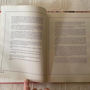 Puede incluir: Libro abierto con cubierta roja, que muestra texto impreso en p&aacute;ginas blancas. Las p&aacute;ginas est&aacute;n abiertas en una doble p&aacute;gina con columnas de texto. El libro est&aacute; sobre una superficie blanca estampada. Un dedo se&ntilde;ala la parte inferior de la p&aacute;gina.