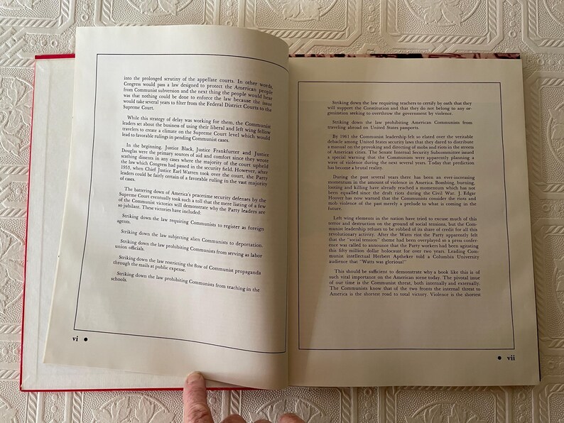 Puede incluir: Libro abierto con cubierta roja, que muestra texto impreso en p&aacute;ginas blancas. Las p&aacute;ginas est&aacute;n abiertas en una doble p&aacute;gina con columnas de texto. El libro est&aacute; sobre una superficie blanca estampada. Un dedo se&ntilde;ala la parte inferior de la p&aacute;gina.