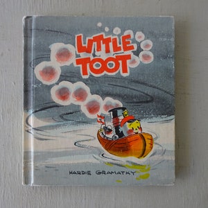 May include: A vintage children's book cover featuring a cartoon illustration of a red and orange tugboat named "Little Toot" with a white smoke trail behind it. The book title "Little Toot" is in red letters on a blue background.