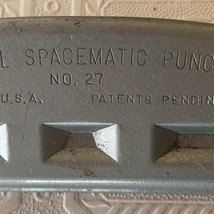 May include: Close-up of a vintage gray metal paper punch. The text "MUTUAL SPACEMATIC PUNCH" and "NO. 27" are visible. The words "MADE IN U.S.A." and "PATENTS PENDING" are also present. The punch has a curved top and three rectangular punch holes.